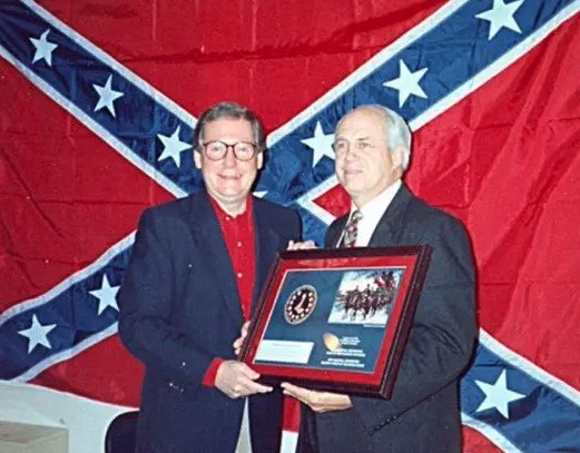 Mitch McConnell with big smile standing next to another white guy accepting an award or something with a giant confederate flag behind them